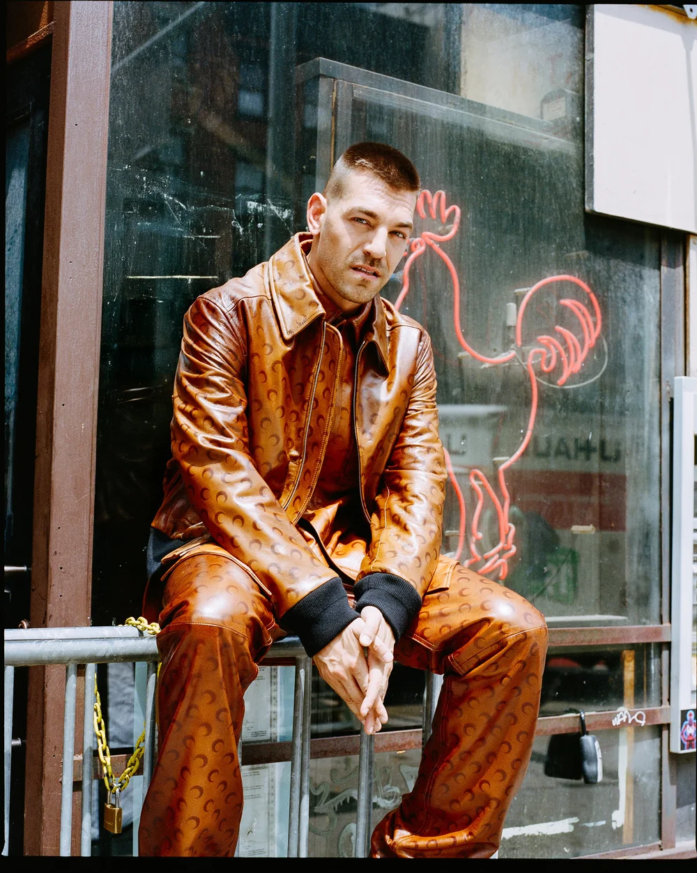 Matt Rogers in a brown leather suit seated by a neon sign for ODDA magazine