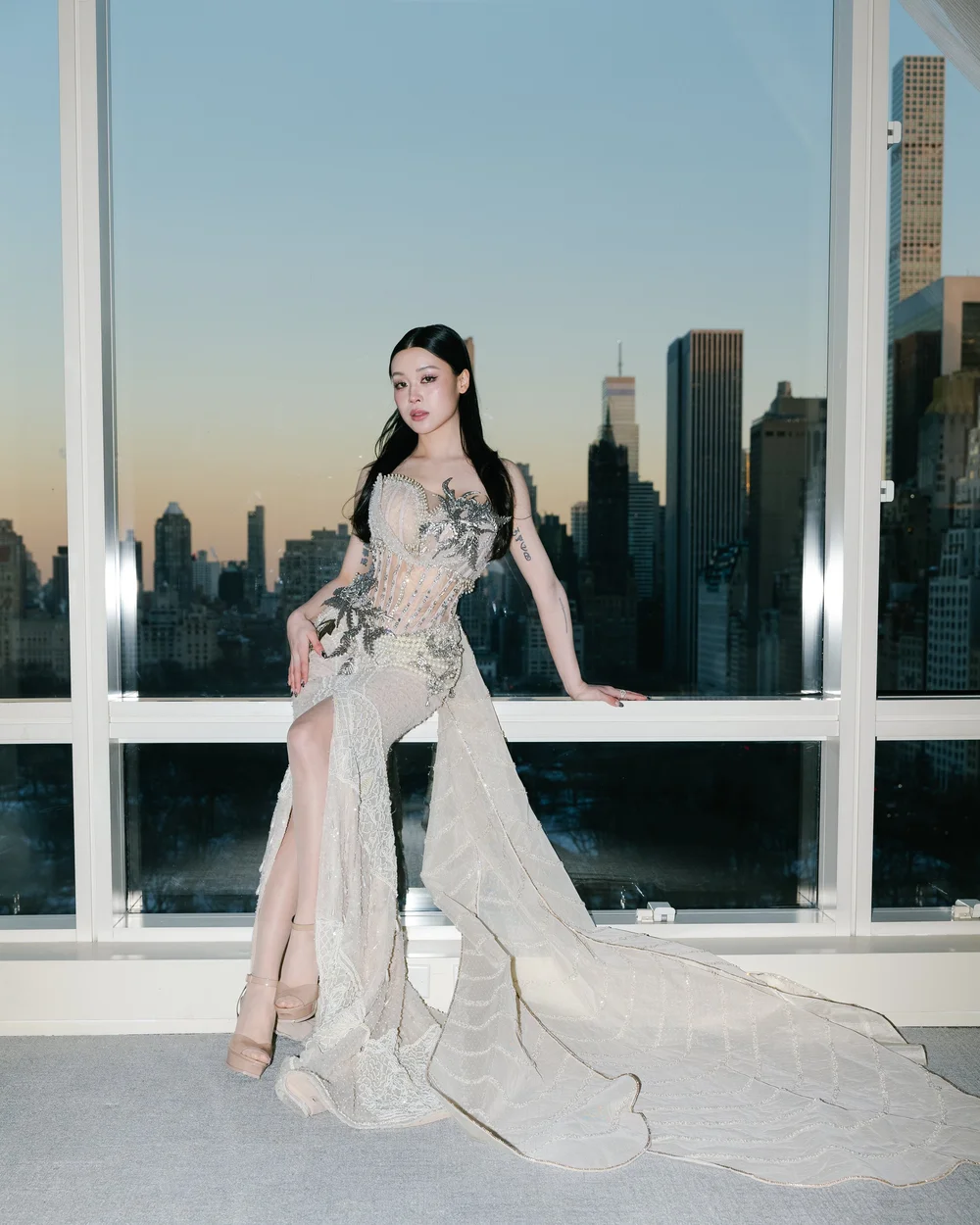Emily Mei in a flowing gown by a window overlooking the Manhattan skyline at sunset